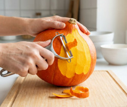 Mains épluchant un potiron orange vif avec un économe sur une planche à découper en bois dans une cuisine claire.