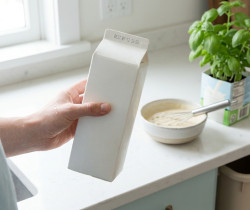 Une personne tenant une brique de lait vierge dans une cuisine lumineuse avec un bol de préparation et du basilic.