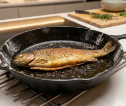 Une truite entière dorée cuite dans une poêle en fonte noire posée sur une grille de cuisine en inox.
